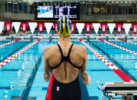 A swimmer wearing a black swimsuit and a blue and yellow swim cap stands at the edge of a pool, preparing for a race. The view is from behind, looking toward the lanes of the pool with red and white lane dividers. In the background, there is a large scoreboard.