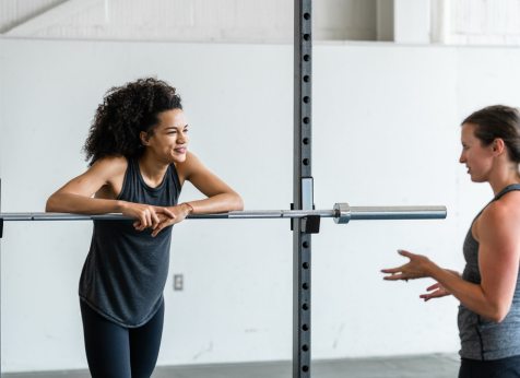 Two women having a friendly conversation in the gym by a squat rack, emphasizing the supportive and motivating environment of strength training and fitness coaching.
